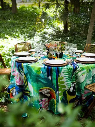 Mesa redonda con mantel tropical de guacamayos, bajoplatos de madera y copas de cristal en un jardín para una boda