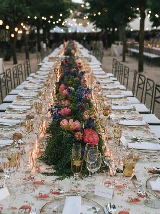 Larga mesa imperial de boda con camino de peonías, delphinium y verde, velas y mantel estampado de flamencos
